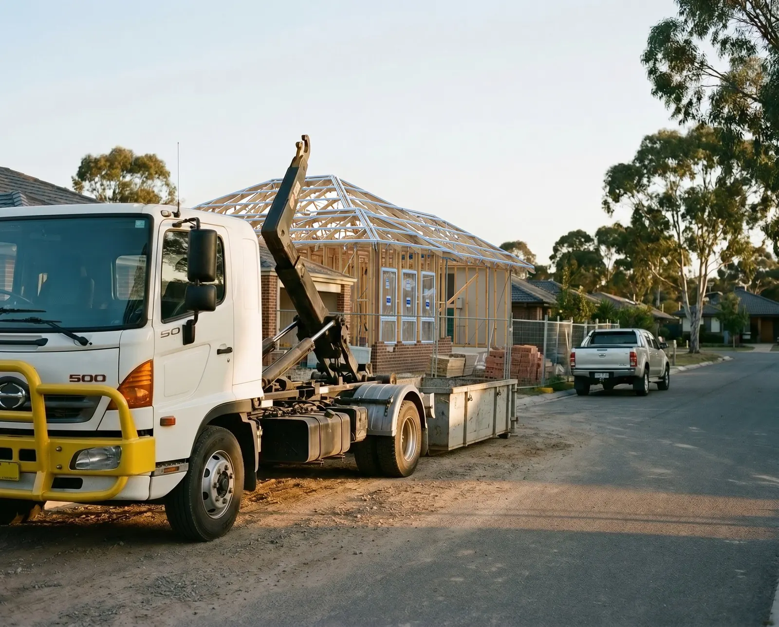 A hook-lift truck parked on a suburban street beside a new house frame at golden hour
